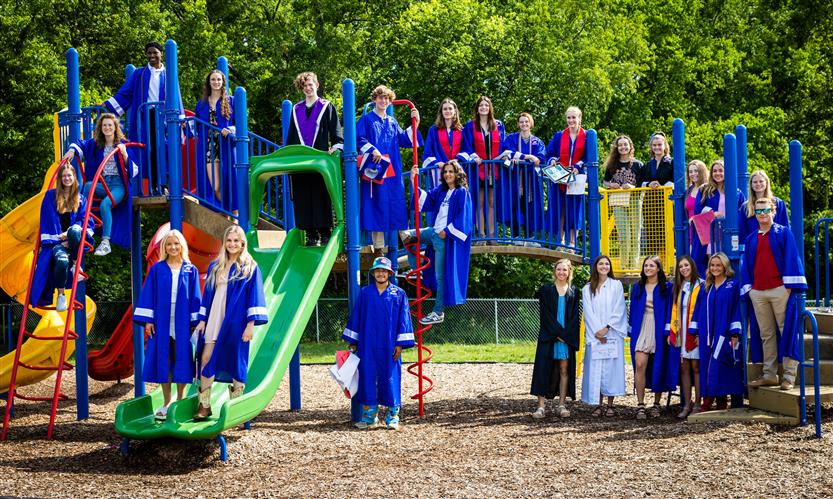 Students wearing graduation robes smile while posing on a playground set